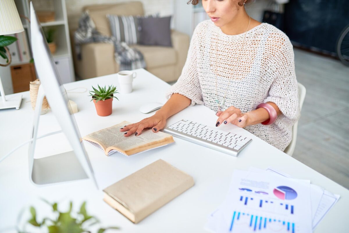 A woman reads a book while working on her computer at home