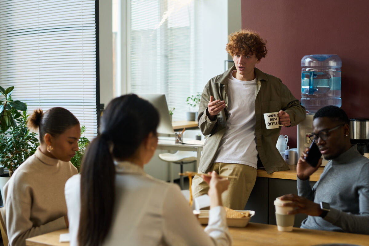 Coworkers gathered in a casual office breakroom setting, chatting and sharing lunch, with one person holding a mug that says “World’s Best Office.”