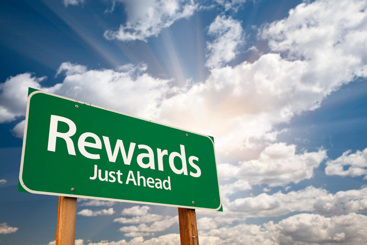 Green road sign reading "Rewards Just Ahead" under a blue sky with clouds